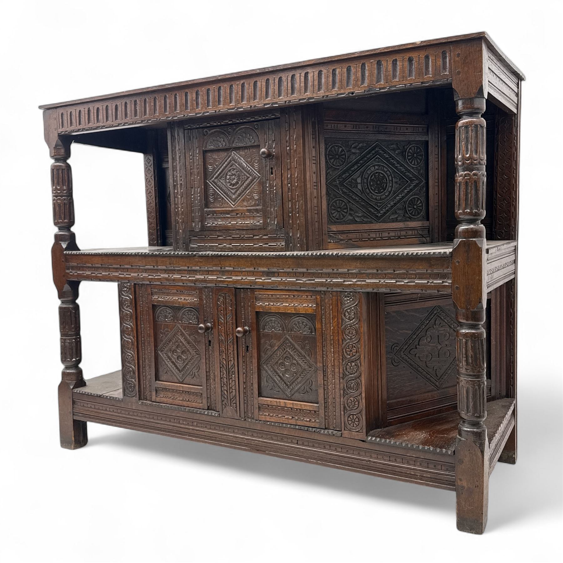 Early 20th-century  oak Jacobean two-tier revival court cupboard, rectangular top with carved notch and stylized foliate carved edge above a carved lozenge pattern frieze and a central cupboard with carved geometric designs and geometric carved panelled sides, above a carved panelled cupboard with two doors of similar geometric design, the whole flanked by two sets of stop-fluted baluster turned columns and supported on stile feet