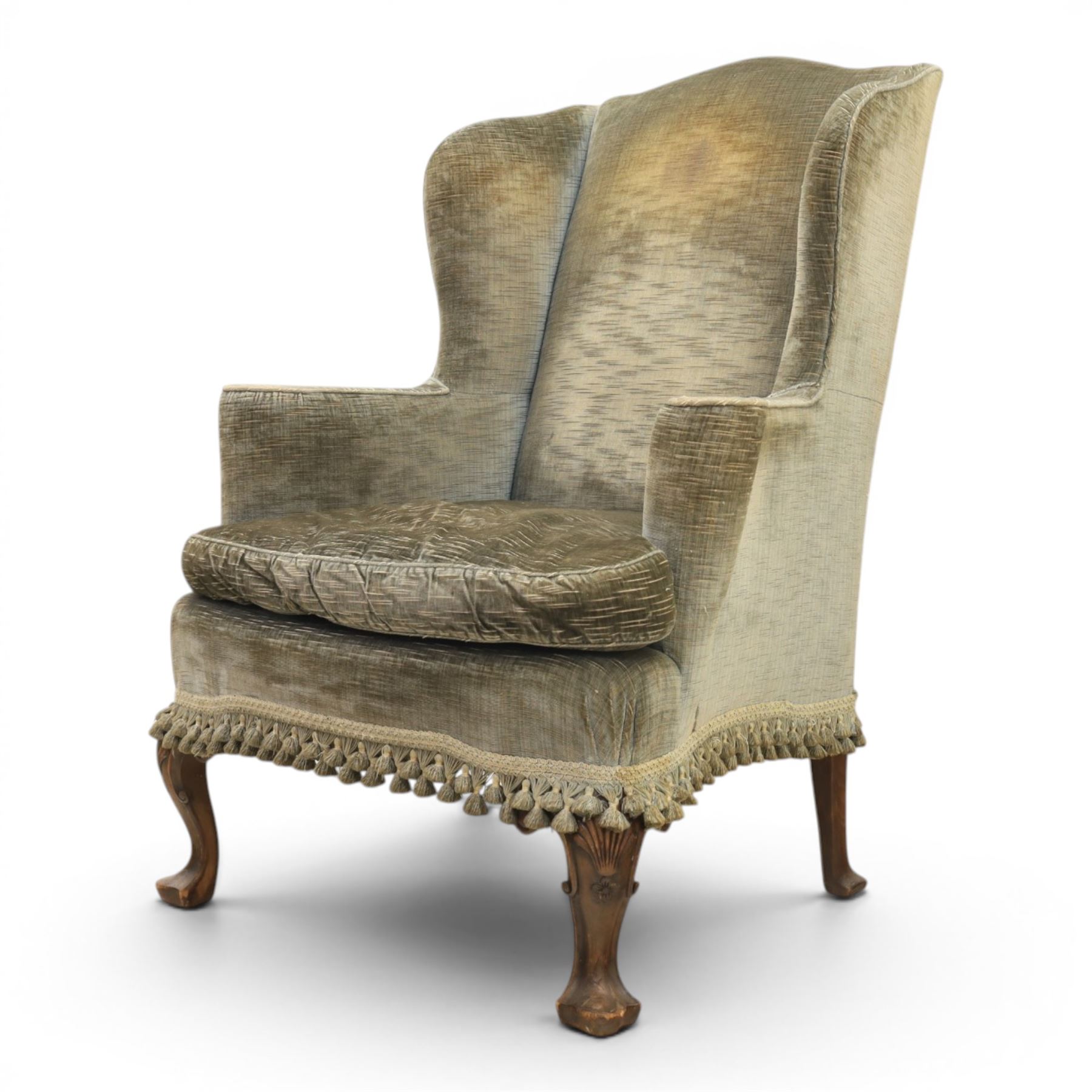 Pair of Georgian style wing armchairs, upholstered in green velvet, each with shaped wing backs, padded scroll arms and loose seat cushions, raised on cabriole supports terminating in pad feet, one with tasselled fringe