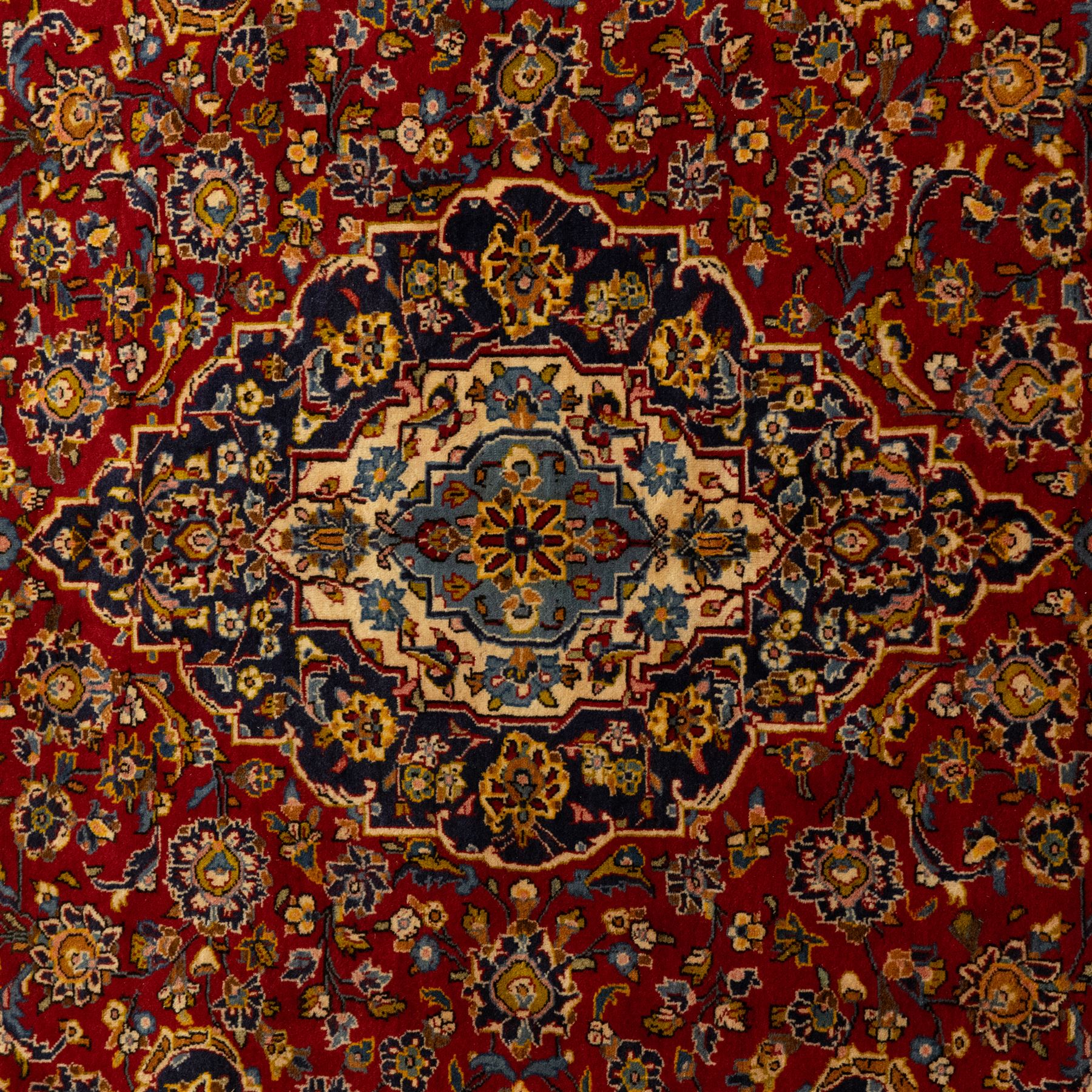 Central Persian Kashan hand-knotted carpet, crimson red field with a central shaped palmette medallion with pendants, filled with dense scrolling and floral motifs, enclosed by a navy blue border decorated with palmettes and meandering vine scrolls, flanked by multiple guard stripes, with cotton fringe ends 