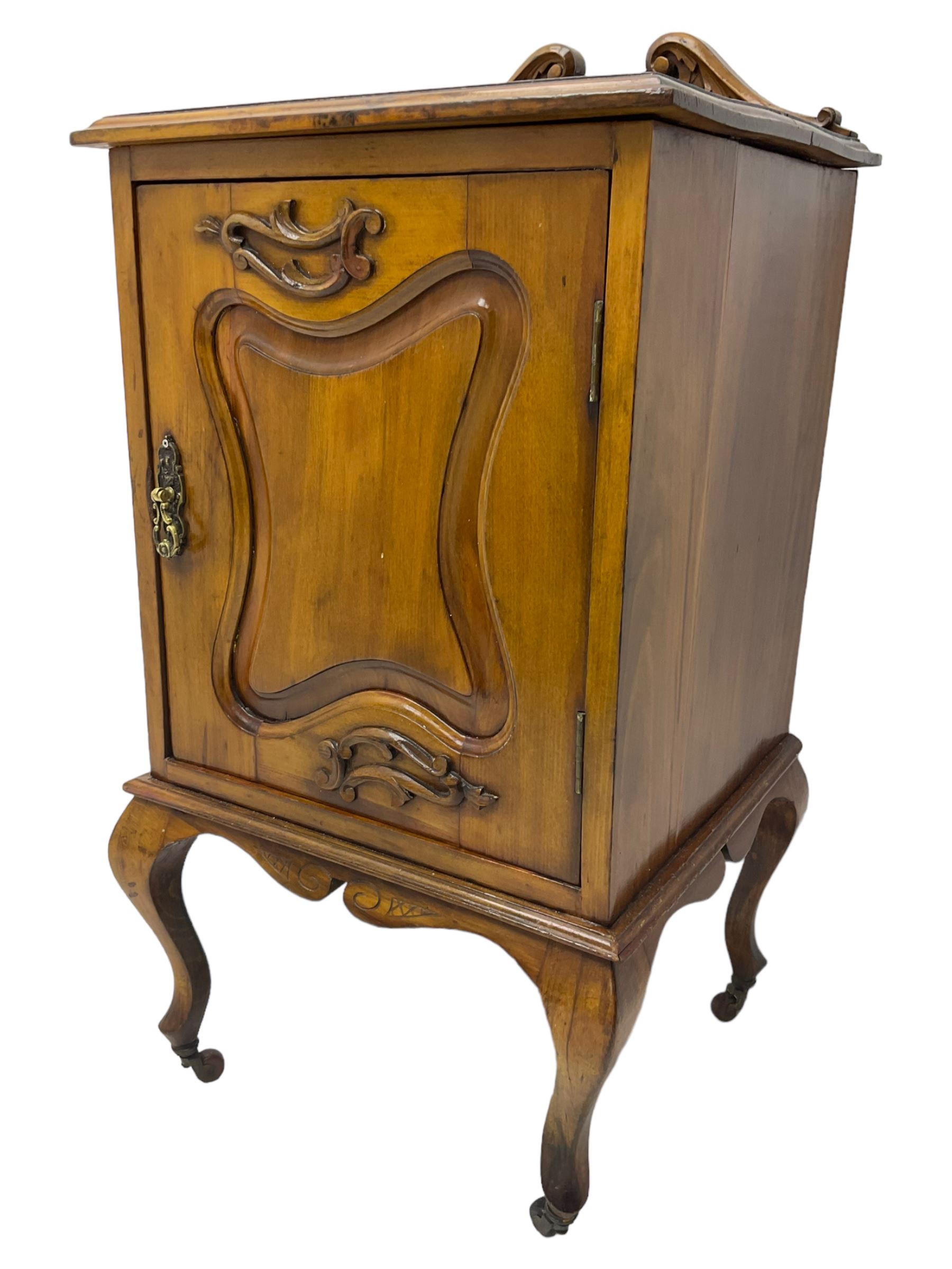 Edwardian Art Nouveau walnut bedside cabinet, raised broken swan neck back with carved flower heads, single fielded panelled door enclosing single shelf, raised on cabriole supports with ceramic castors