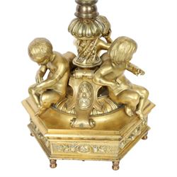 Second Empire style gilt metal table lamp, the hexagonal plinth base with applied foliate decoration, upon six turned feet, supporting three seated allegorical putti, the first modelled with a tablet and stylus representing literature, the second with hammer representing sculpture, and the third with Corinthian column capital representing Architecture, each intersected by a stylised scroll with central flower head panel, mounted upon an onyx and gilt metal Corinthian column with wrythen flower moulded and part fluted base, supporting a cream octagonal shade, overall H86cm
