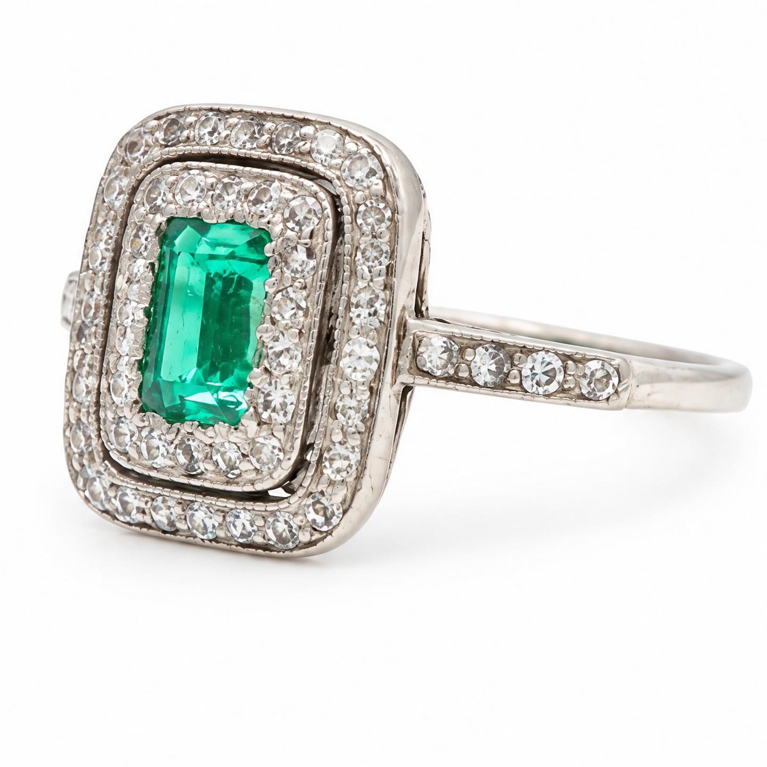 Early 20th century palladium emerald and diamond halo cluster ring, single rectangular cut emerald of approx 0.30 carat, surrounded by two rows of milgrain set diamonds and diamond set shoulders