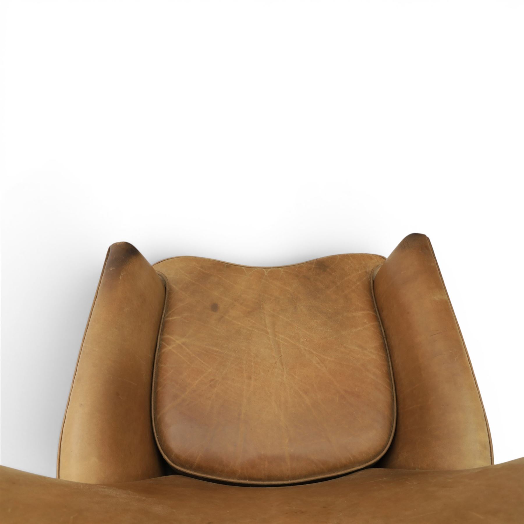 20th century tan leather armchair, the curved wing back above low square arms, shaped loose seat cushion with piped edge, close-nailed side detail, raised on concealed castors