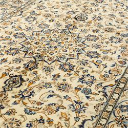 Central Persian Kashan hand-knotted rug, ivory ground, with a small central floral medallion and pendants, the field decorated with fine blue, ochre and beige scrolling vines, flowerheads and palmettes, within an ivory border of repeating floral sprays and meandering vines, flanked by narrow guard stripes, with cotton fringes