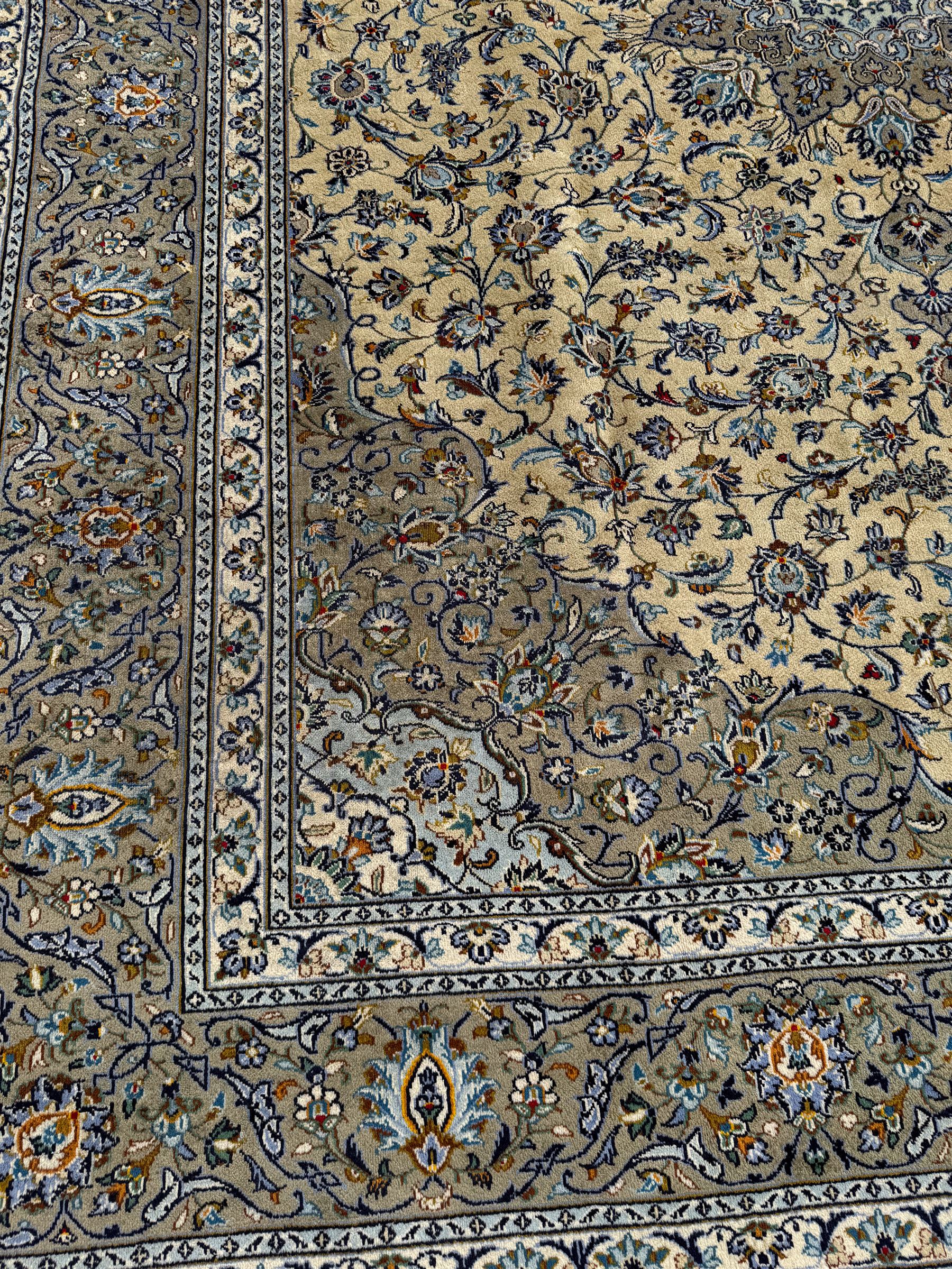 Central Persian Kashan pale khaki and ivory ground carpet, shaped central medallion enclosing small stylised motifs, the field decorated profusely with trailing foliate branches and plant motifs, guarded border with repeating floral design