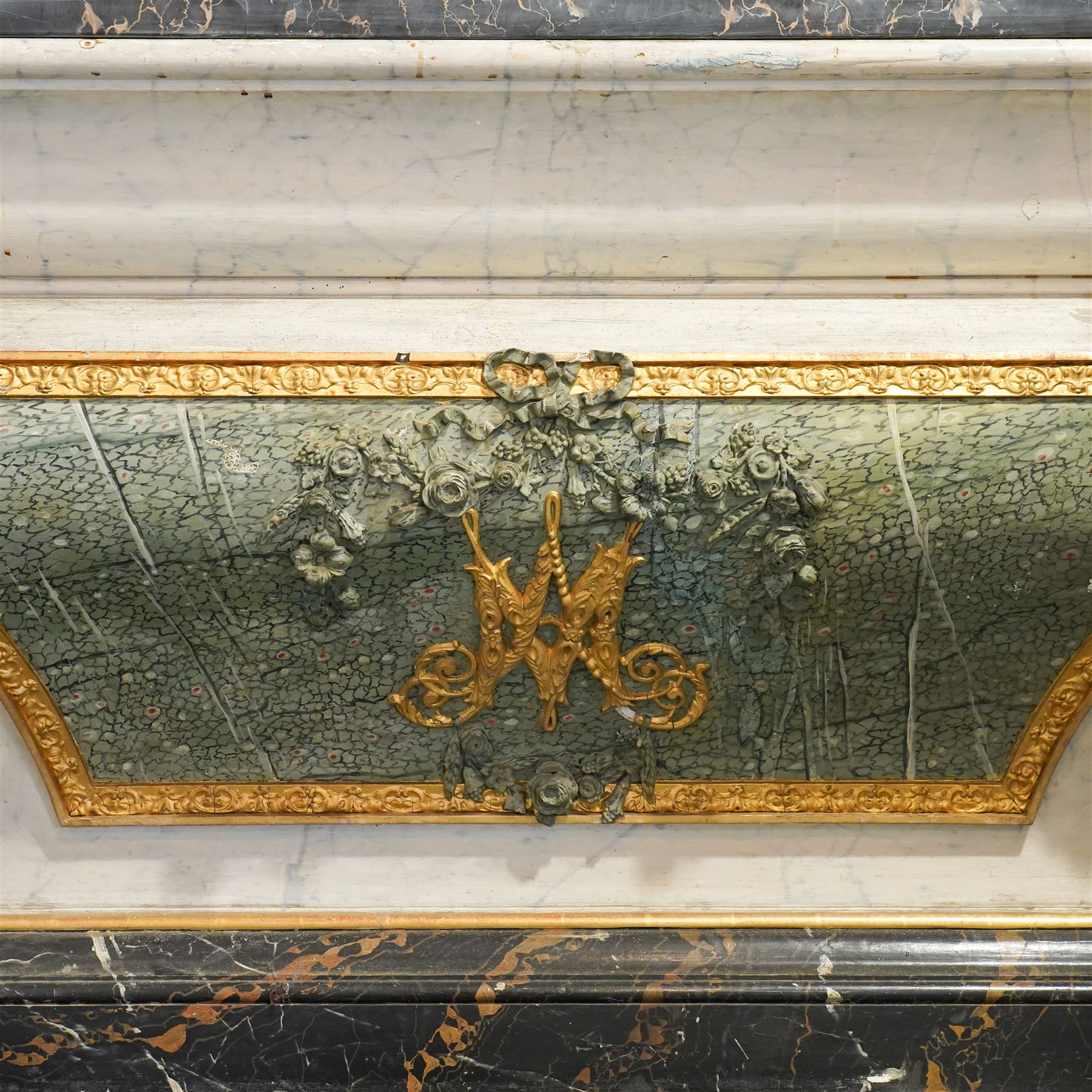 A 20th century Italian Neoclassical console table of architectural pedestal form, inspired by Roman architectural prototypes, with marble top and simulated marble scagliola-type finish; the rectangular marble top with grey veining within a black and yellow marble border, above a pedestal base finished in a finely executed faux-marble surface and enriched with parcel-gilt scrolling acanthus terminals, floral garlands, a central ribbon-tied motif in relief, and further raised flowerheads and a central gilt monogram, terminating in a stepped plinth base