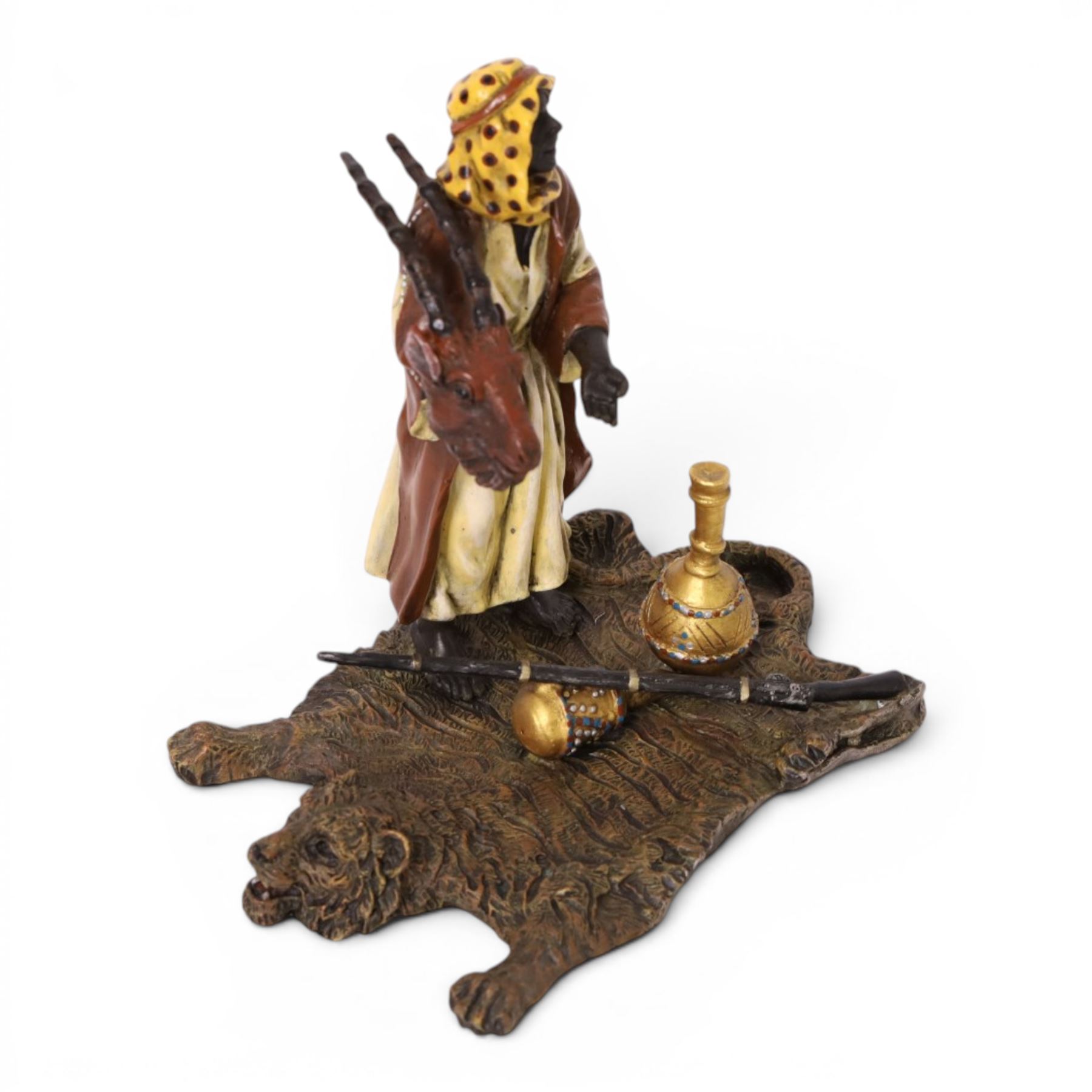 After Bergman - bronze Arab trader holding an antelope head while standing on a tiger skin, with a rifle and jars at his feet, H14cm
