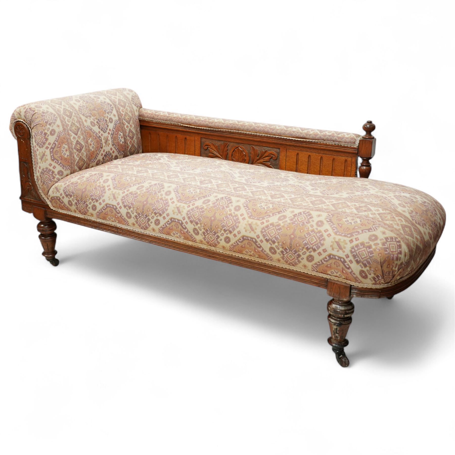 Late Victorian walnut chaise longue, upholstered in geometric patterned fabric, the scroll end and padded back above carved side panel with foliate and roundel motifs, shaped seat rail on turned front supports with ceramic castors