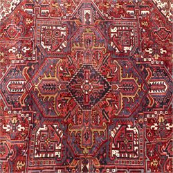 North West Persian Heriz hand-knotted carpet, madder red field with central bold angular steeped medallion with serrated outlines, flanked by stylized palmettes, hooked leaves and geometric motifs, spandrels similarly decorated within a wide geometric border and multiple guard borders in tones of madder red, indigo blue, ivory and olive green, with cotton fringe ends  