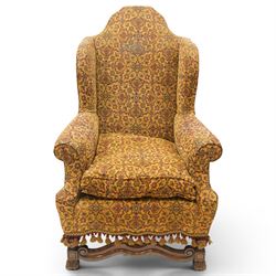 Pair of William & Mary design wingback armchairs, the arched high-back over rolled arms with a loose seat cushion and sprung seat, upholstered in old-gold ground foliate patterned fabric, raise on scroll feet united by a front serpentine stretcher