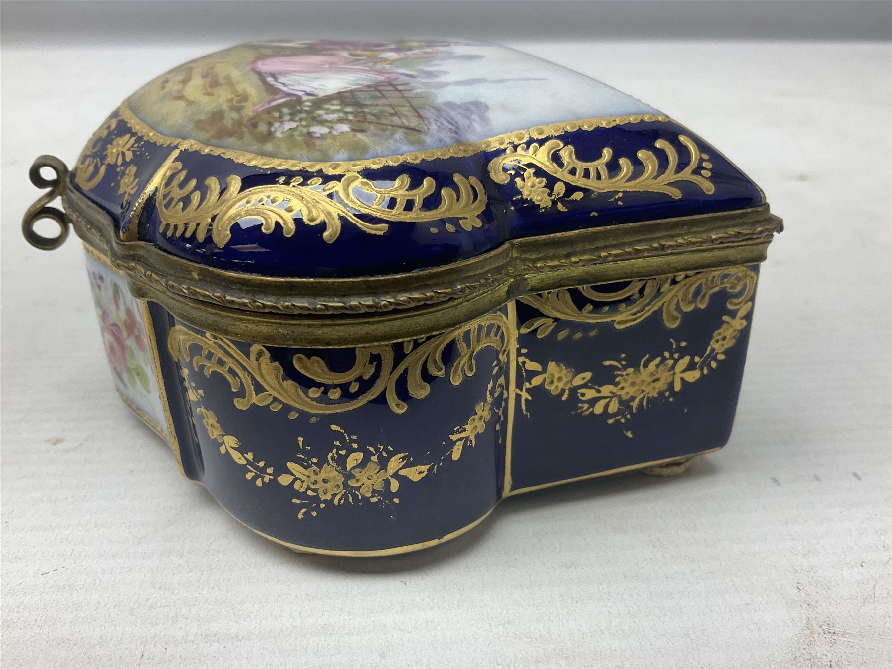 Sevres style late 19th/early 20th century trinket box decorated in the rococo style, the shaped cover painted with an 18th century courting couple in a garden scene opening to reveal the interior painted with floral sprays, the conforming body with a painted panel of floral decoration, all reserved upon cobalt blue ground with foliate gilt borders, with interlaced L's mark beneath, L12cm H6cm