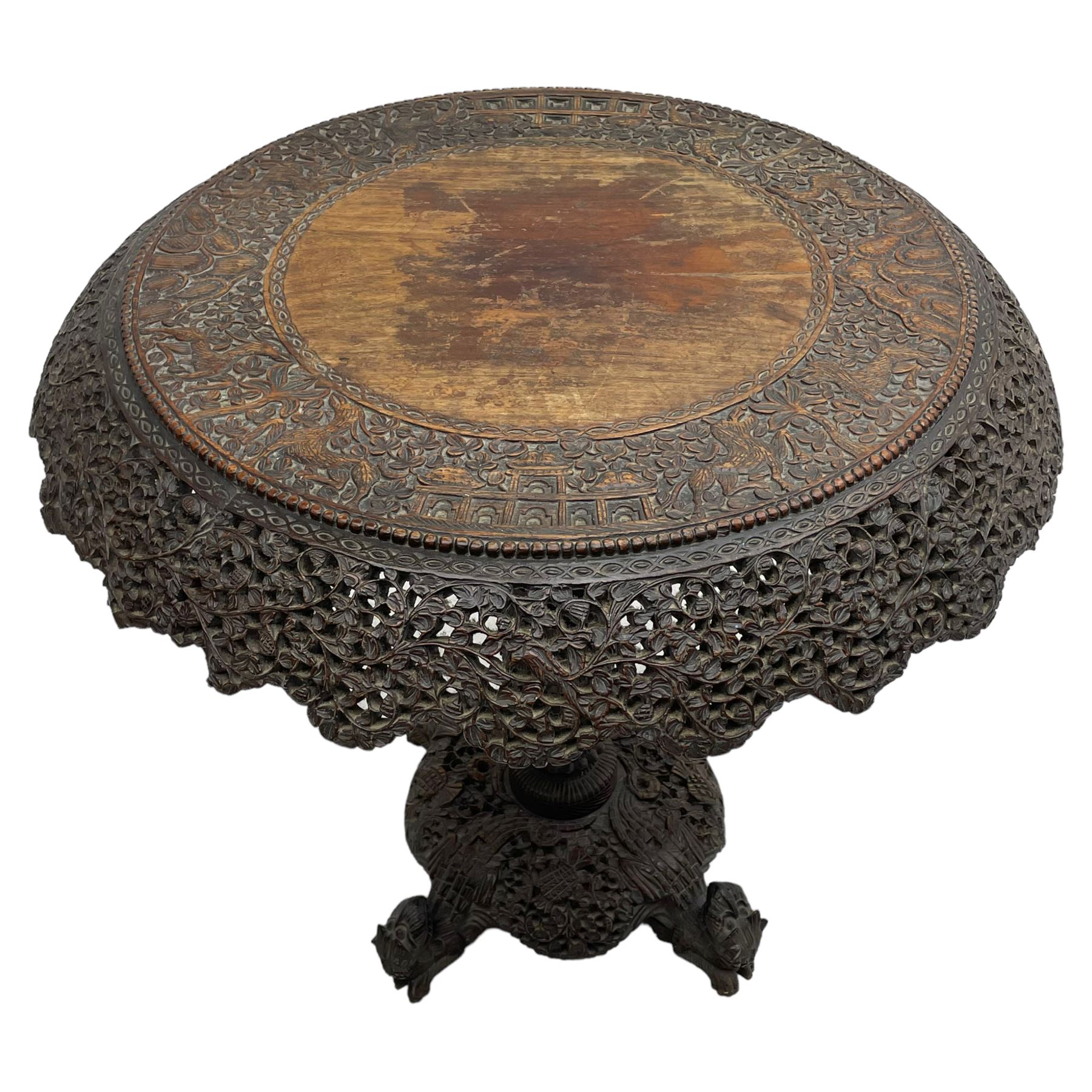 Late 19th century Anglo-Indian carved hardwood occasional table, circular tilt-top carved profusely with mythical animals and foliage decoration, pierced and carved with interlaced leafy branches, leaf carved pedestal supports on a raised platform with further carved and pierced foliage decoration, four carved feet in the form of mythical beasts with gaping jaws 