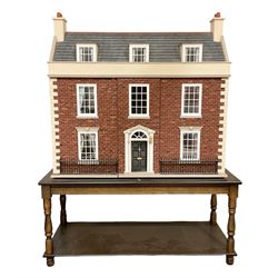 Good quality late 20th century wooden dolls house by M. James dated 1992, as a 19th century style three-storey double fronted property with textured brick walls and faux slated pitched roof with dormer windows, the double hinged front elevation with faux iron railings, opening to reveal four rooms on two floors with central hall stairs and landing; the roof hinged along the ridge opening to give access to two further rooms with central bathroom; windows to both front and rear elevations with drapes; simulated floor boards and tiling throughout with decorated walls, coving and ceilings; wired for electric lighting with bank of switches on the back; bears makers plaque W92cm H89cm D47cm; on modern oak two-tier coffee table as stand.
Auctioneers Note: the WC cistern is no longer fitted in the bathroom and is not included with this lot.