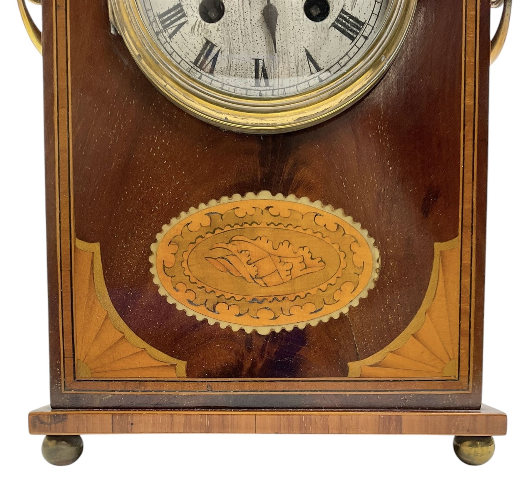 Edwardian- 8-day mantle clock in a mahogany case with a serpentine top and contrasting inlayed panels in satinwood and boxwood to the front, on a rectangular plinth raised on ball feet, silvered dial with Roman numerals and steel spade hands, twin train Parisian rack striking movement, sounding the hours and half hours on a coiled gong. With pendulum.