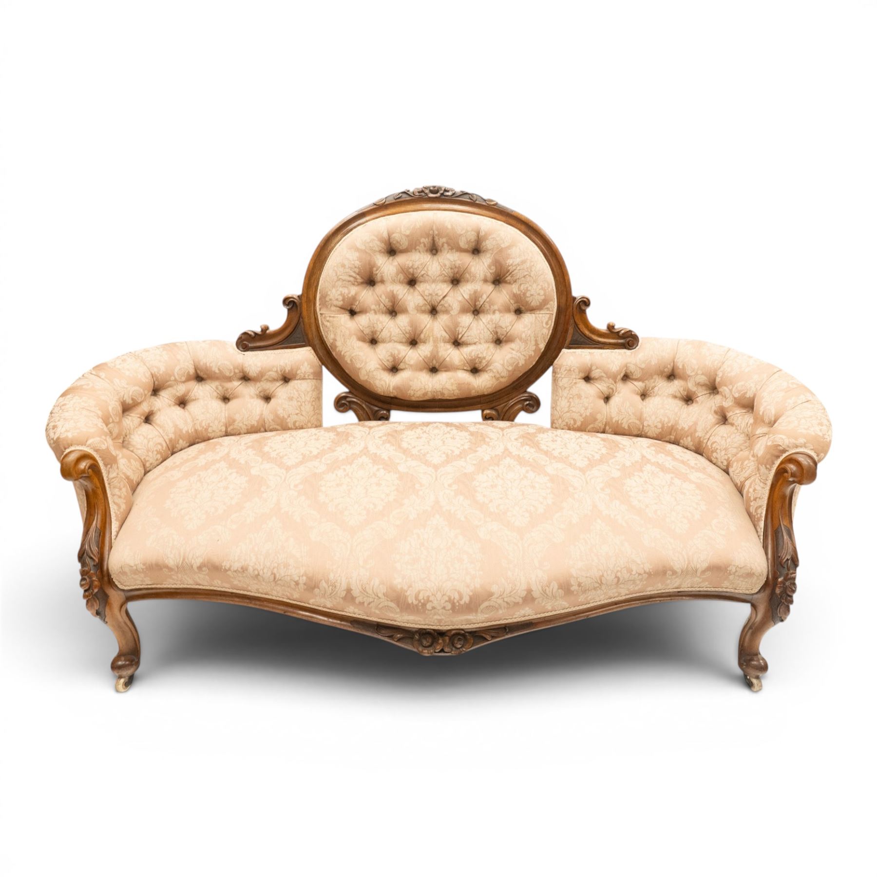 Victorian carved walnut framed double-ended settee with serpentine front, the central oval cameo back with flowerhead carved cresting rail, flanked by foliate C-scroll spandrels, the scroll carved arm terminals over floral carved cabriole supports with scroll feet and ceramic castors, the buttoned back and sprung seat upholstered in pale pink foliate patterned damask fabric