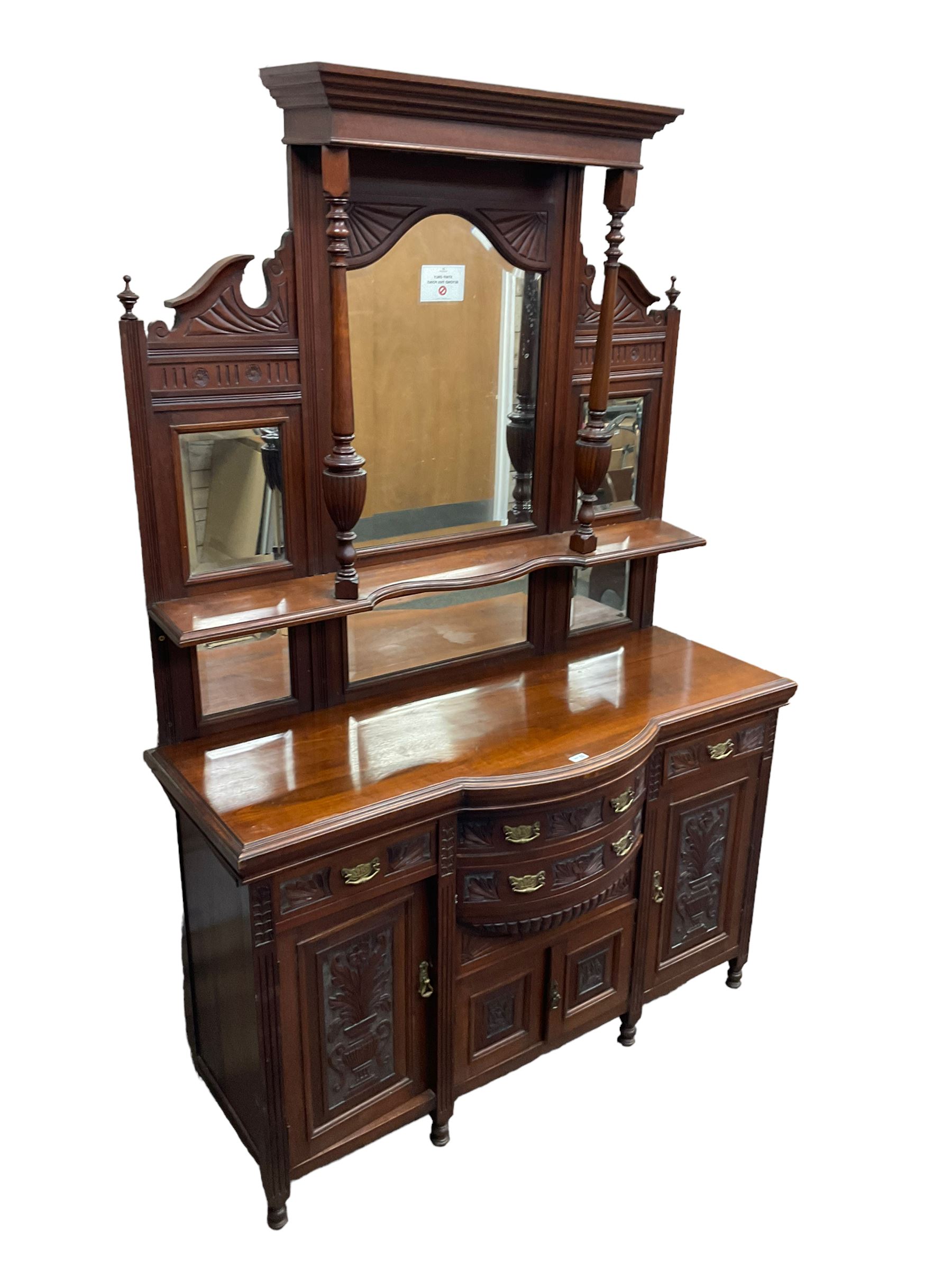 Edwardian walnut sideboard, projecting cornice supported by turned supports, the back fitted with three bevelled mirrors beneath fluted and flowerhead carved friezes, the lower section with four drawers and four panelled cupboard doors carved with foliate and urn designs, fluted uprights on turned feet
