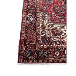 Persian Heriz red ground rug, central scrolled medallion on dark ground decorated with floral motifs, the surrounding field decorated with stylised plant motifs and smaller geometric symbols, guarded border with repeating pattern punctuated with rosettes and flower heads 