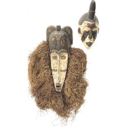 Two African carved masks, comprising of a white painted mask with carved facial details and a long form mask with carved eyes and open mouth surrounded with raffia decoration, longest example L60cm