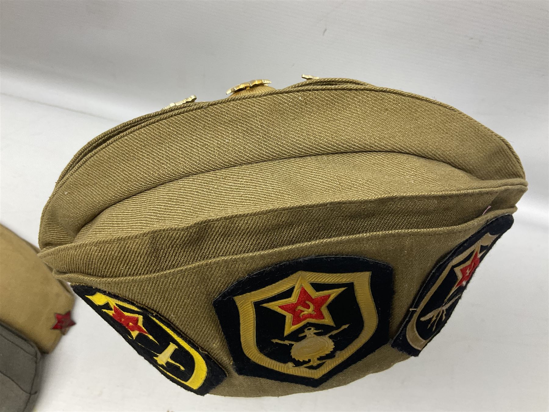 Four side caps - WW2 Russian, post-WW2 Russian and two East German; all with badges, one with multiple metal badges (4)