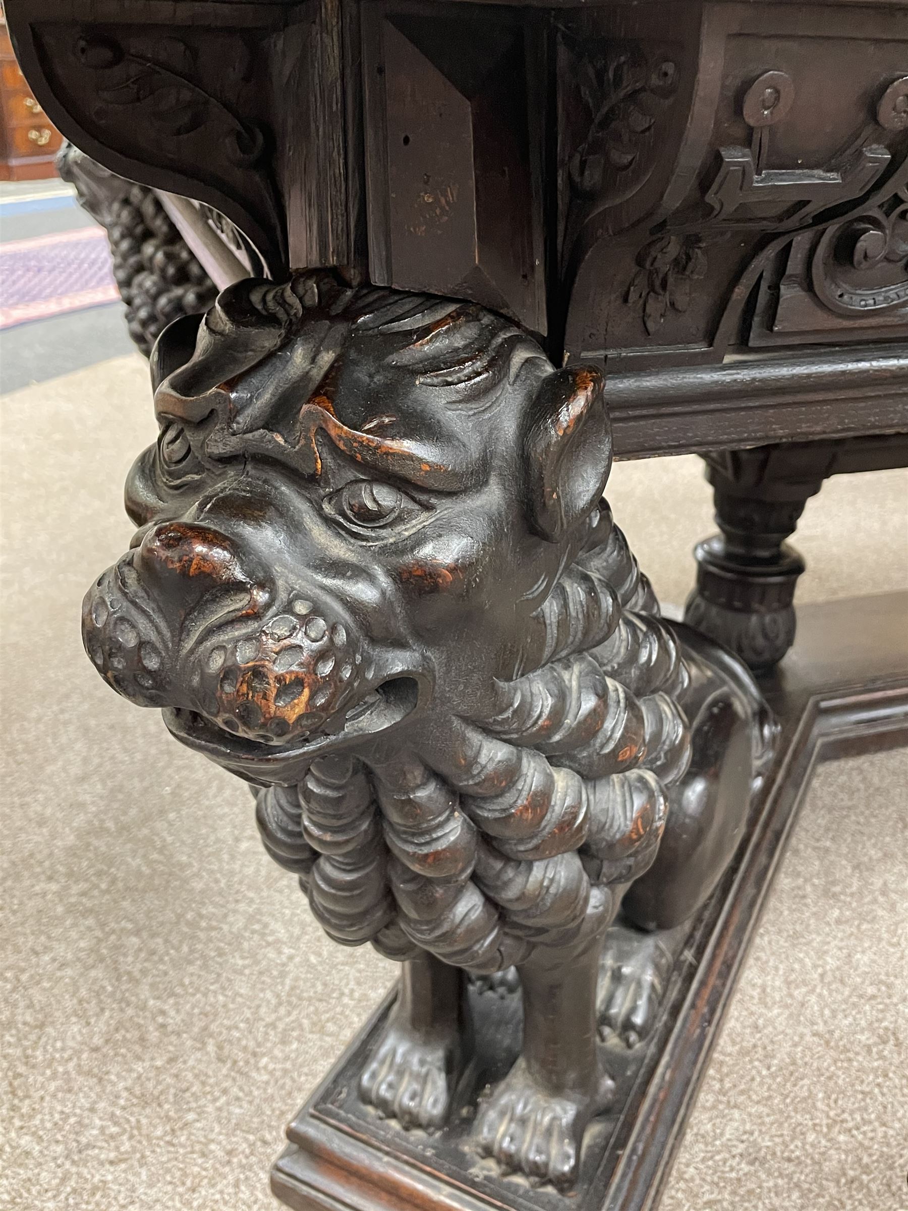 Large 19th century carved oak library table, the rectangular top with lunette carved edge over cartouche panelled and carved  scroll frieze, the central tapering rectangular support relief carved with masks and garlands, on four carved seated lion supports at each corner, on flat angular stretcher with recessed castors stamped ‘Bartholomew & Fletcher, London’