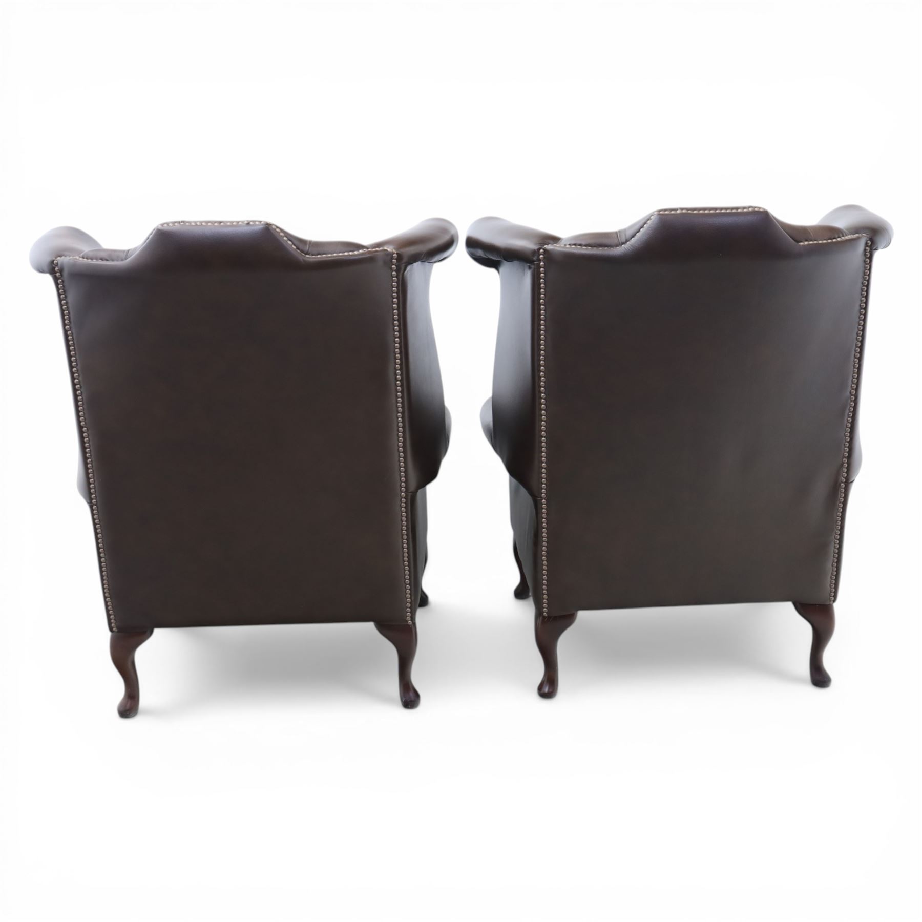 20th century Chesterfield style brown leather wingback armchairs, with button upholstered backs and wings, shaped scroll arms with studded borders, loose seat cushions, raised on cabriole supports with pad feet