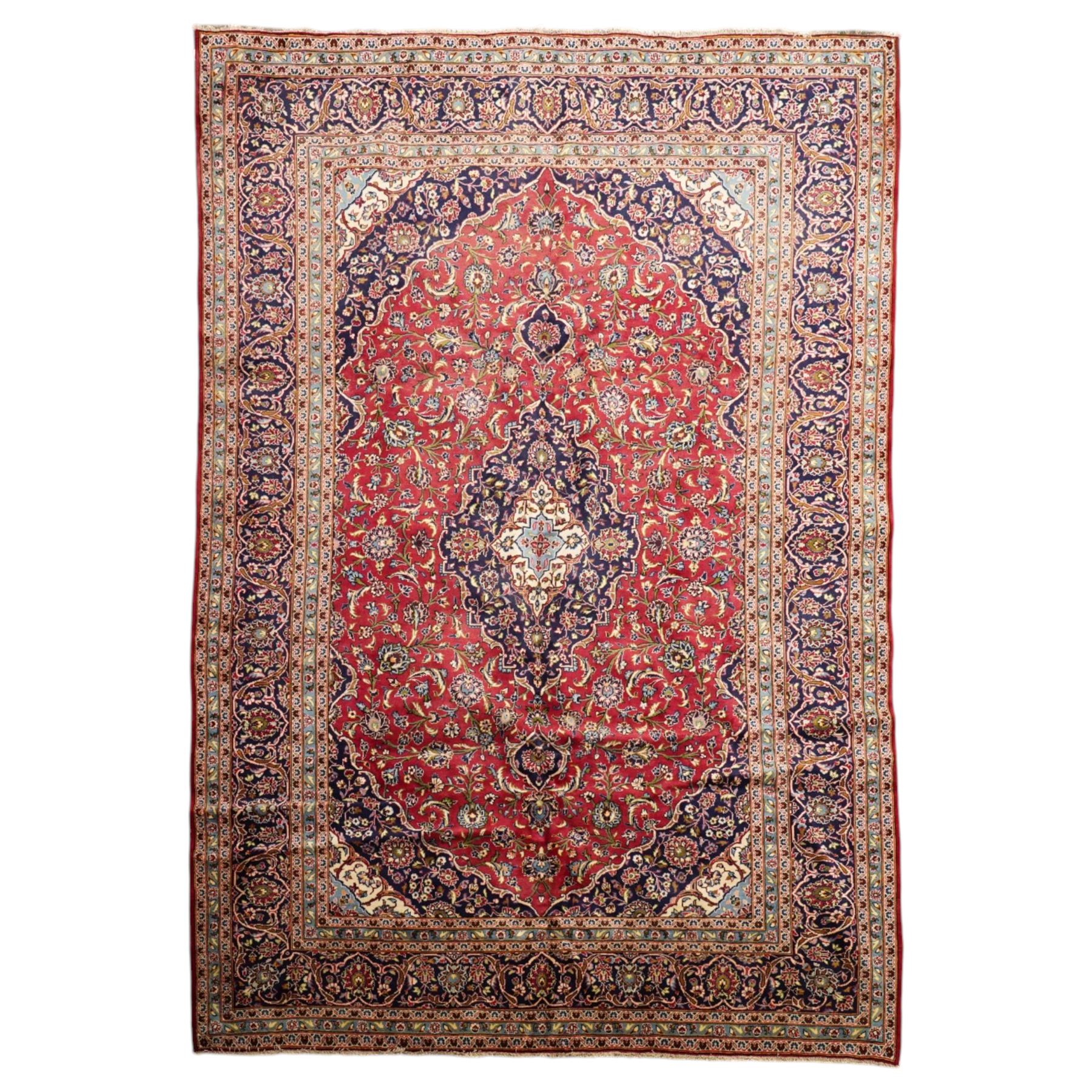 Persian Kashan hand-knotted carpet, madder red ground centred by shaped dark navy and ivory floral medallion with pendants, surrounded by scrolling vines, palmettes and flowerheads, dark navy spandrels, enclosed by dark navy floral border with multiple narrow floral guard stripes