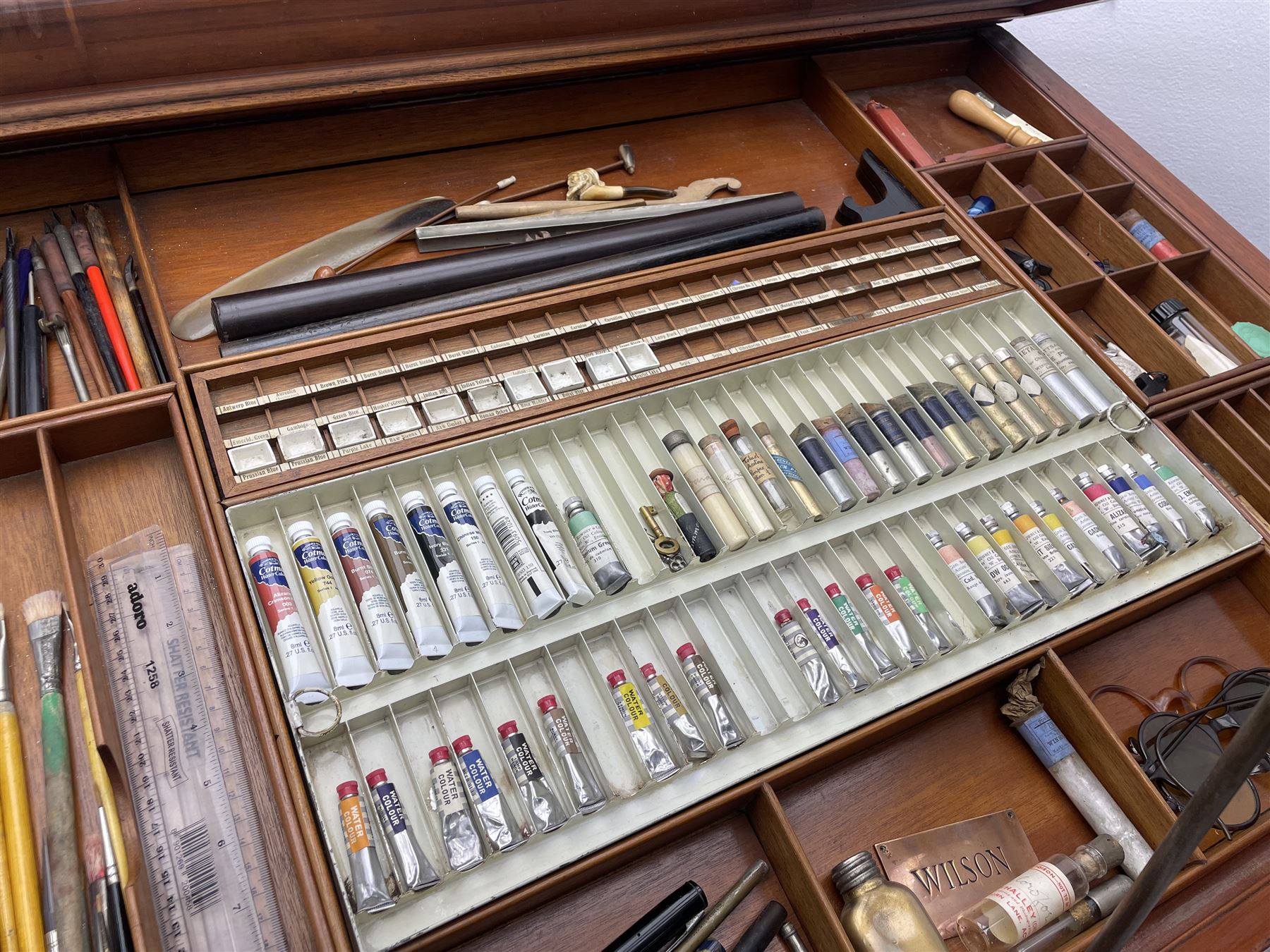 Edwardian mahogany artist's cabinet once belonging to the British artist Eli Marsden Wilson (1877-1965), raised panelled back mounted by carved brackets, sloped and hinged glazed lid enclosing paint holders and compartments, up-and-over panelled door revealing three slides, fitted with two banks of five graduating drawers with recessed brass handles, on skirted base.

Together with a quantity of artists' equipment and tools including paints, paintbrushes, charcoal, chalks, etc. and various prints relating to the life of the artist, including Quaker anti-war posters, Victorian Stevengraphs, and various chromolithographs 

Provenance: Removed from the studio of Eli Marsden Wilson. The cabinet comes to us for sale through family descent. The vendor is descendant of Marsden. Many further etchings removed from the cabinet are being sold in the Spring Art Sale, Friday 15th March 2024, Lots 265-271.

Notes: born in Ossett, Eli Marsden Wilson was the only son of Alfred Wilson, a foreman beamer, and Emma Marsden, and had five sisters. After studying at Wakefield College of Art, he moved to the Royal College of Art in London, where he became a pupil of Sir Frank Short. He had his first exhibit at the Royal Academy in 1905, the same year he married fellow artist Hilda Mary Pemberton.

A Quaker, vegetarian, and pacifist, he was a conscientious objector in World War I and as such was imprisoned in 1917 for two years. In September 1922 he was commissioned by Princess Marie Louise to produce miniature etchings for Queen Mary's Dolls' House at Windsor Castle; two such etchings are being sold in the Spring Art Sale, Friday 15th March 2024, Lot 265.