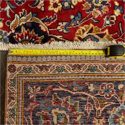 Central Persian Kashan hand-knotted carpet, crimson red central field with central floral medallion with floral vine-work and palmettes, within dense floral spandrels and a wide navy blue border decorated with continuous foliate motifs and multiple guard borders, with cotton fringe ends  