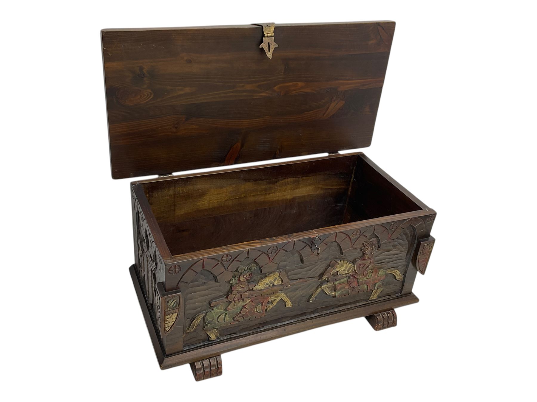 Mid-20th century carved hardwood chest, the rectangular hinged top with decorative strap hinges, the front and sides deeply carved with medieval-style jousting knights, heraldic shields, and architectural elements, raised on shaped block feet