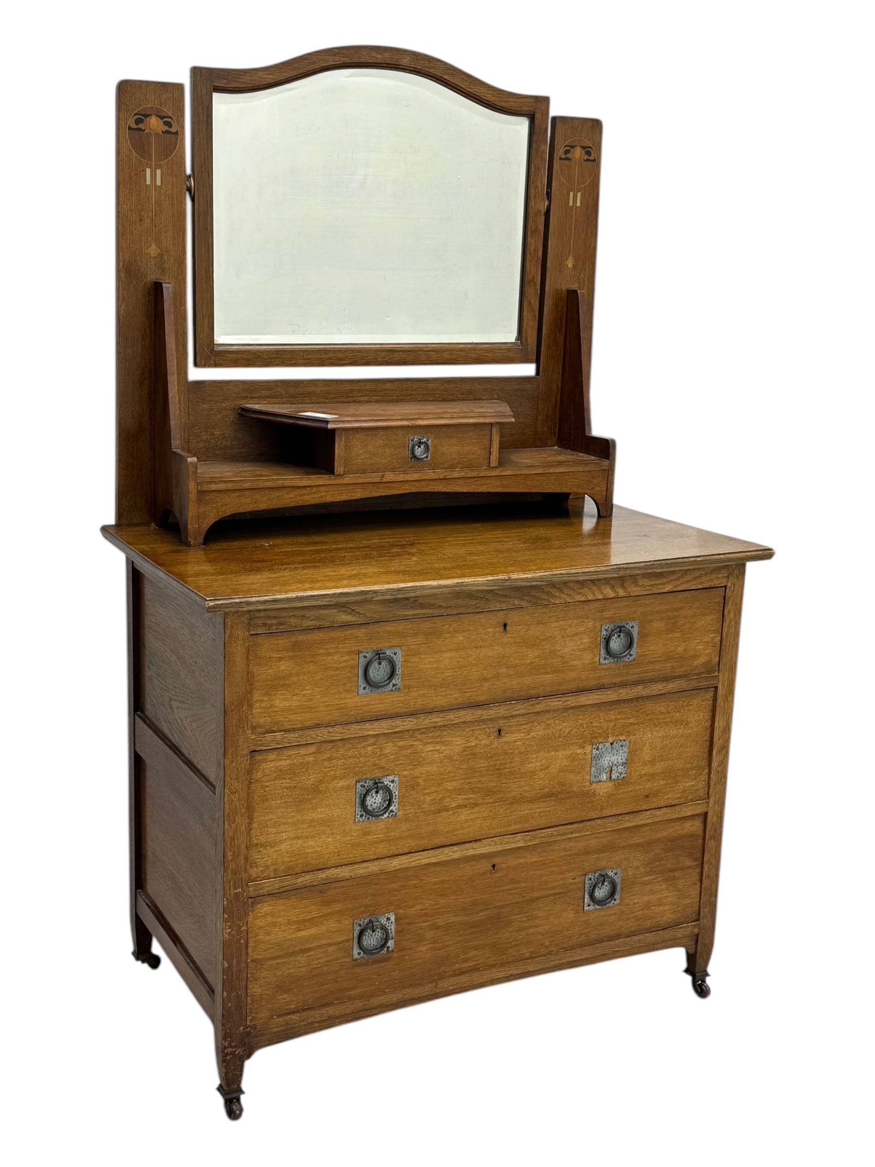 Early 20th century Arts and Crafts oak dressing chest, the arched mirror flanked by floral marquetry inlaid uprights above a shelf and drawer, over three long drawers with square hammered metal plates and circular pulls, on slender supports with castors