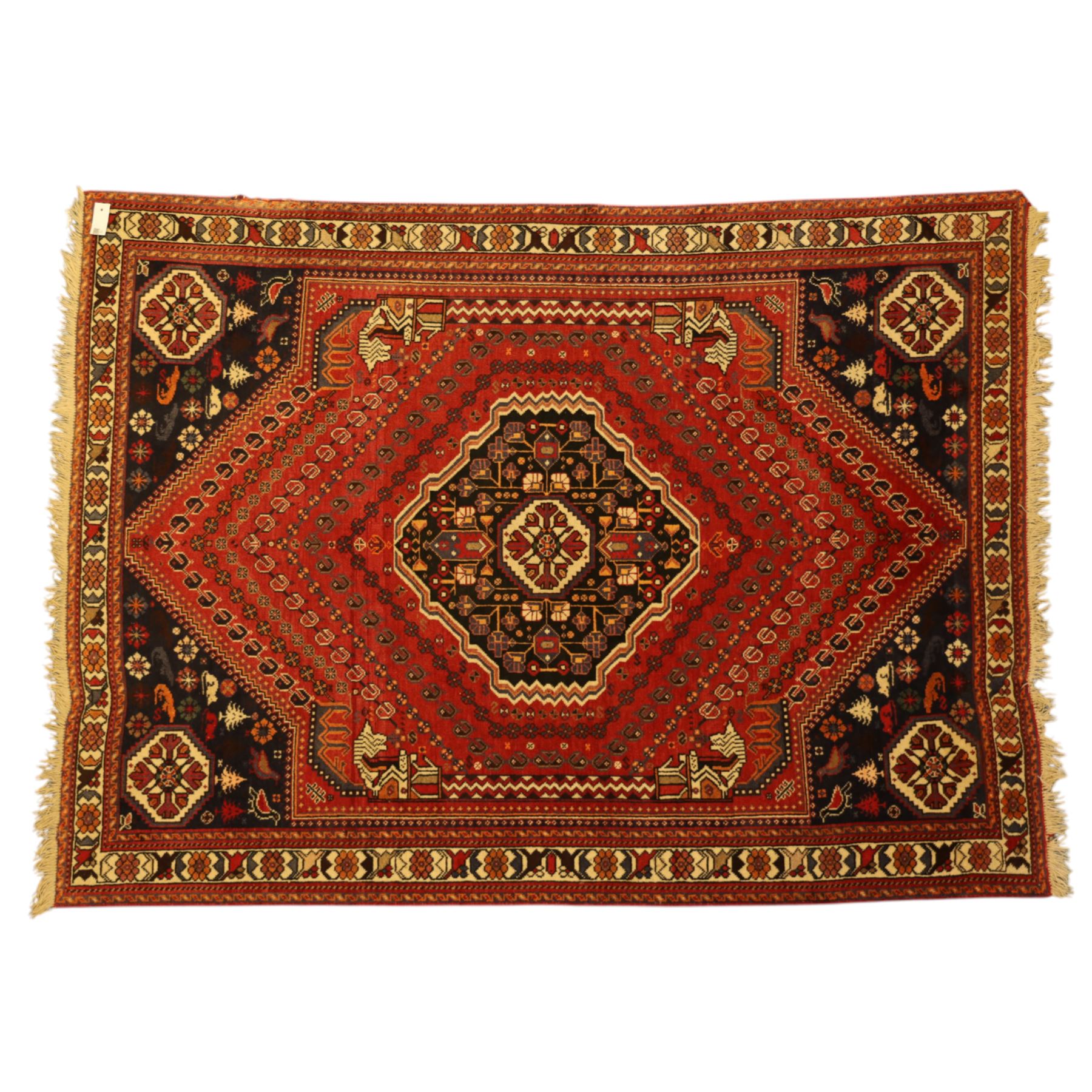 Persian Shiraz rug, hand-knotted tribal rug, bold geometric central medallion, set within a large hexagonal crimson field with angular stepped corners in dark blue, with boteh motifs, rosettes, and other tribal symbols arranged in diagonal rows, the corners with indigo spandrels decorated with zoomorphic and arboreal motifs, main border composed of a floral meander motif with large rosette, flanked guard borders with vine and leaf motifs 