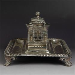 George IV silver desk stand, of rectangular form with rounded corners and oblique gadrooned rim, upon four embossed pad feet, with central pedestal dish of part fluted form with domed lid engraved with crest and with bud finial, with two recesses either side housing faceted glass inkwells, each with silver collars, hallmarked John & Thomas Settle, Sheffield 1825 & 1827, H11cm