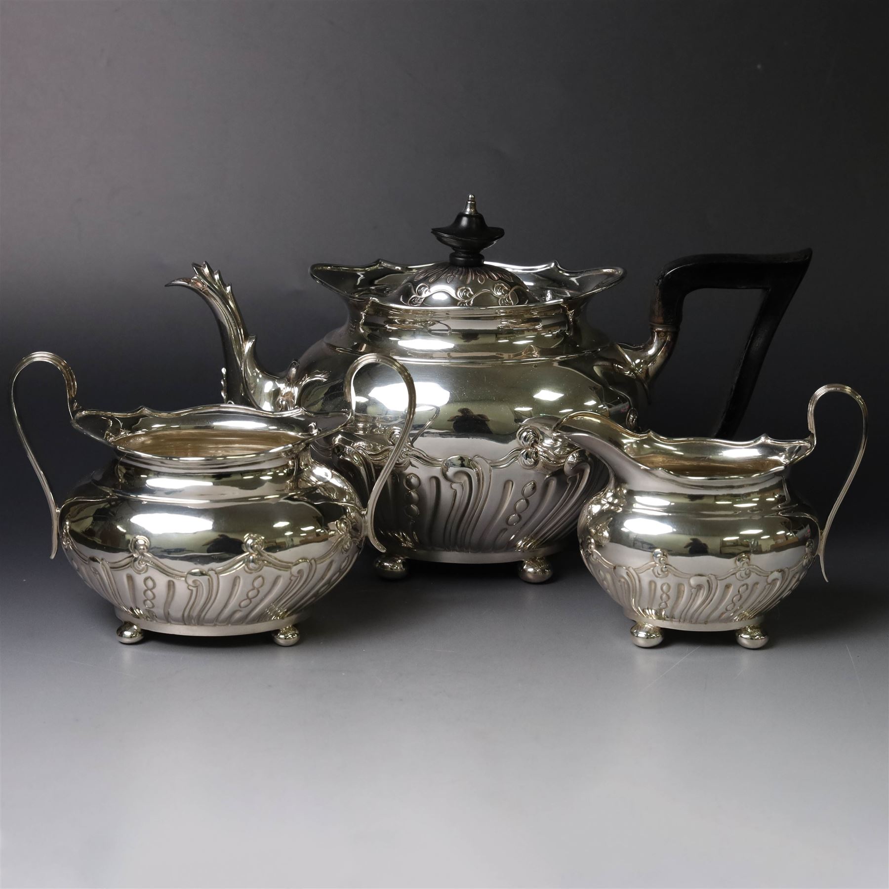 Edwardian silver three piece tea service, comprising teapot, twin handled open sucrier and a cream jug, each of oval form with embossed scrolling decoration and upon four bun feet, the teapot with ebonised wooden handle and finial, hallmarked Thomas Bradbury & Sons Ltd, Sheffield 1903, teapot H16cm
