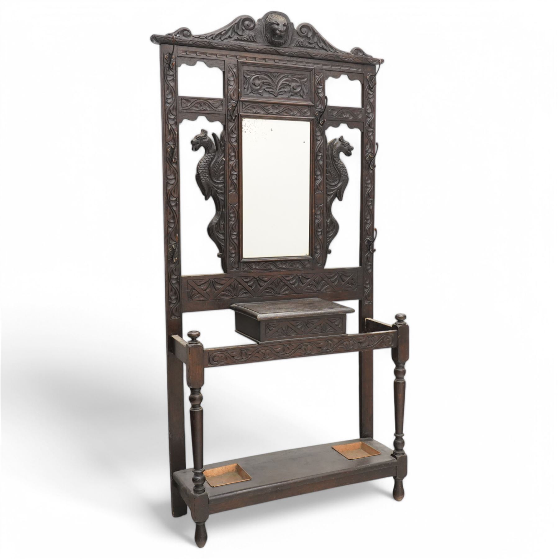 Late Victorian heavily carved oak hall stand, the scrollwork pediment with carved lion's head, bevelled mirror plate flanked by carved dragon motifs, over hinged storage compartment and umbrella stands with drip trays, on turned front supports