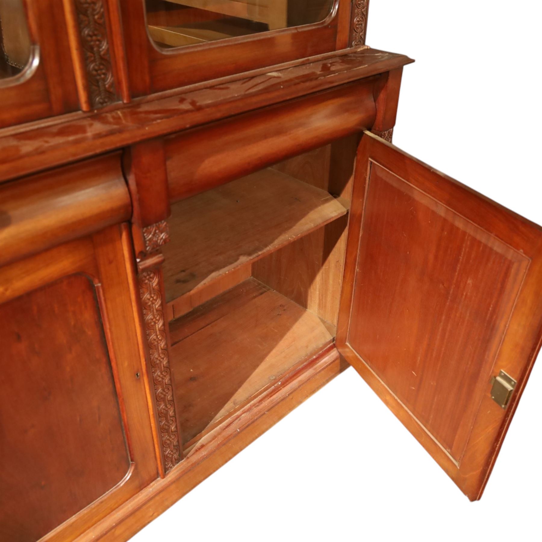 Victorian carved mahogany enclosed bookcase on cupboard, projecting moulded cornice over twin glazed doors enclosing adjustable shelves, flanked by foliate carved uprights, the lower section fitted with two cushion moulded drawers over twin panelled cupboard doors, divided and flanked by conforming carved uprights, on plinth base