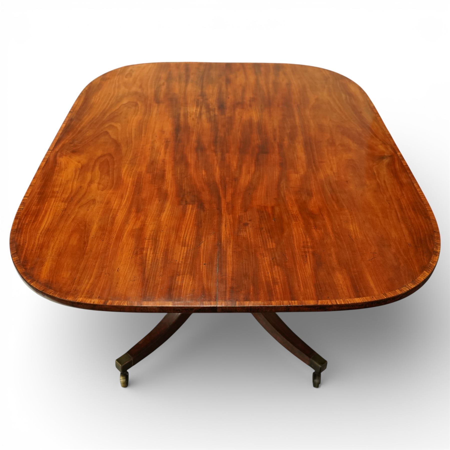 Regency figured mahogany breakfast table, figured rectangular tilt-top with rounded corners and rosewood banding, on turned column with four out-splayed supports with brass cups and castors 