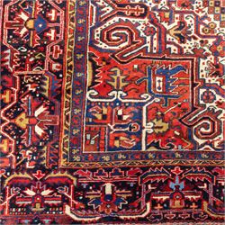 North West Persian Heriz hand-knotted carpet, madder red field, with a large angular stepped central medallion with serrated outlines, flanked by bold stylized palmettes, hooked leaves and geometric devices, spandrels similarly decorated within a wide geometric main border featuring angular floral motifs, enclosed by multiple guard borders in madder red, indigo blue, ivory, olive green and soft ochre, with cotton fringe ends 