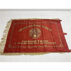 1970s Soviet banner printed in gold on a red ground, roughly translates as 'Transferable Banner - To the winner in the Socialist Competition' and 'Work Study Live by Communism' verso; wreath of wheat ears to either side; tassels on three sides 110 x 155cm