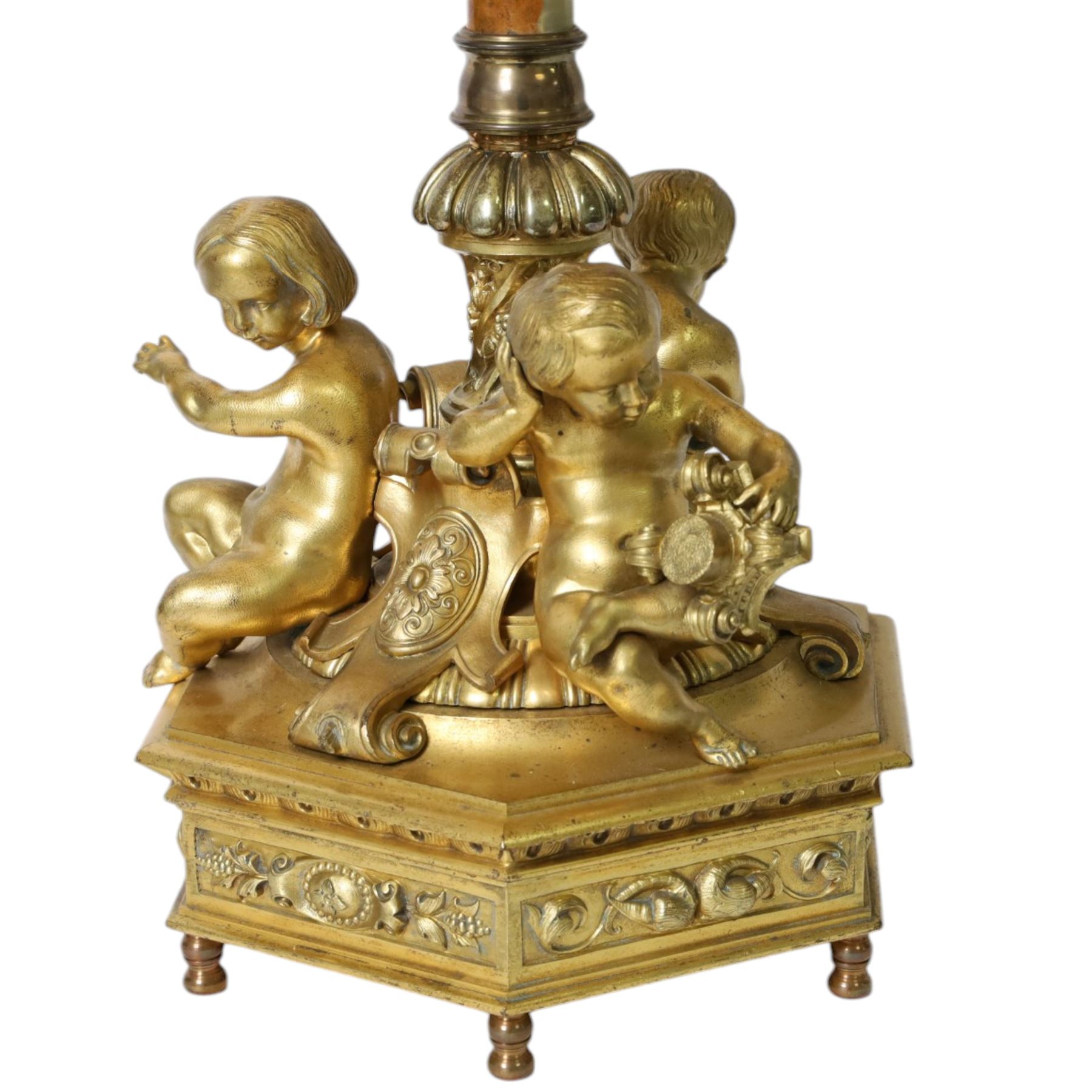 Second Empire style gilt metal table lamp, the hexagonal plinth base with applied foliate decoration, upon six turned feet, supporting three seated allegorical putti, the first modelled with a tablet and stylus representing literature, the second with hammer representing sculpture, and the third with Corinthian column capital representing Architecture, each intersected by a stylised scroll with central flower head panel, mounted upon an onyx and gilt metal Corinthian column with wrythen flower moulded and part fluted base, supporting a cream octagonal shade, overall H86cm
