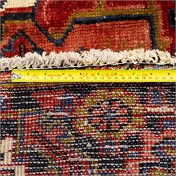 North West Persian Heriz hand-knotted carpet, madder red ground, with a large central geometric medallion with pendants, surrounded by angular foliate and floral motifs incorporating stylised rosettes, palmettes and serrated vine work, enclosed by ivory spandrels and a dark navy border with repeating geometric flowerheads and angular meandering vines, flanked by narrow patterned guard stripes, with cotton fringes