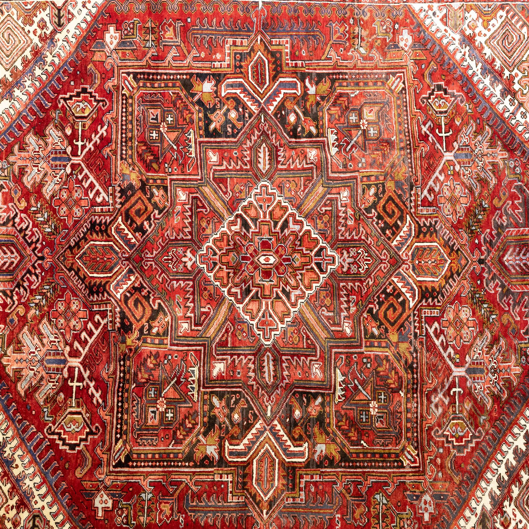 North West Persian Heriz hand-knotted carpet, madder red field, with a large angular stepped central medallion with serrated outlines, flanked by bold stylized palmettes, hooked leaves and geometric devices, spandrels similarly decorated within a wide geometric main border featuring angular floral motifs, enclosed by multiple guard borders in madder red, indigo blue, ivory, olive green and soft ochre, with cotton fringe ends 