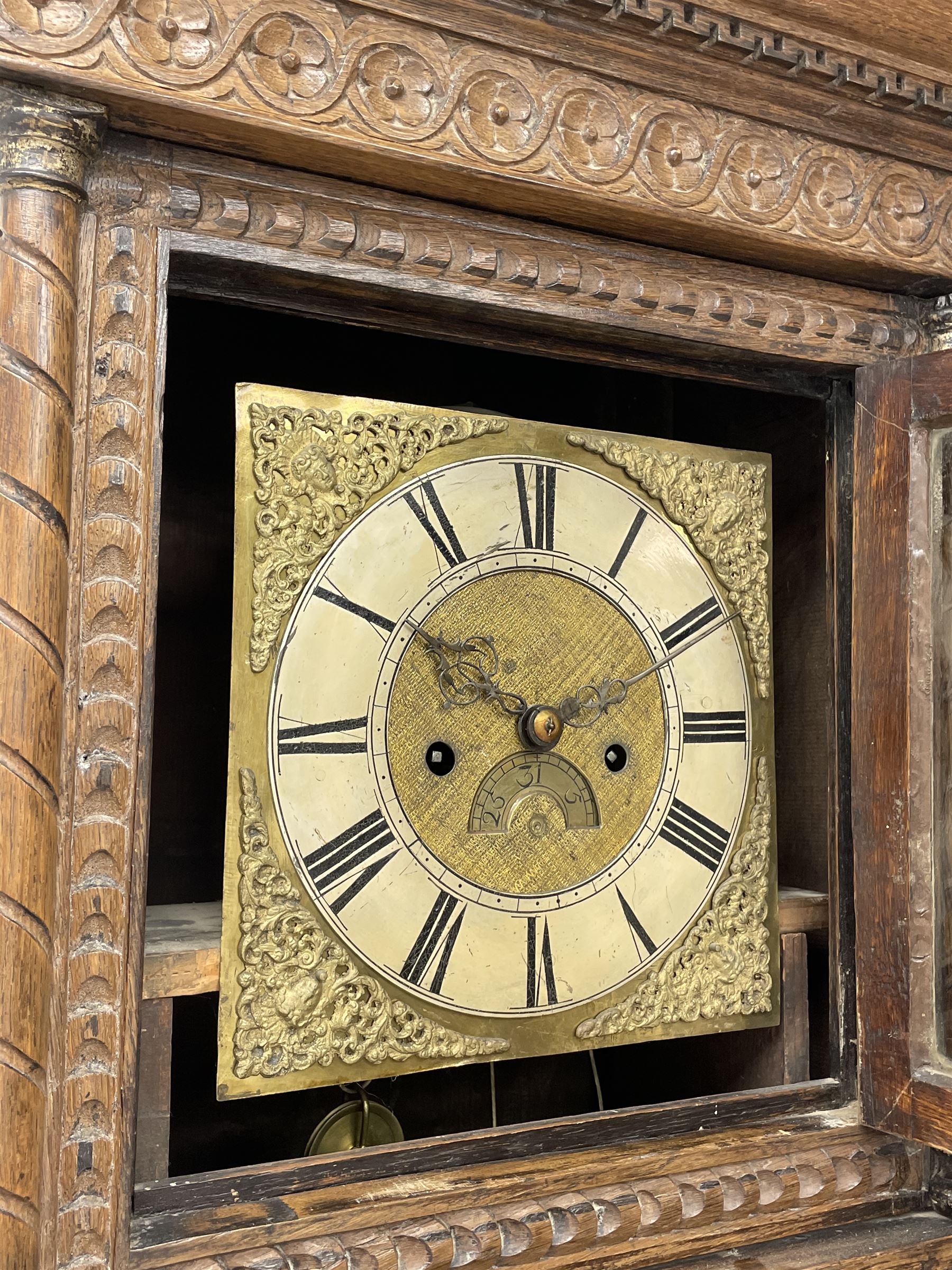 19th century carved oak longcase clock, projecting cornice over guilloche carved frieze, square brass dial with silvered Roman chapter ring and calendar aperture, decorated with ornate mask cast spandrels, the trunk door and base carved with potted plant, twin train driven eight day movement striking on bell
