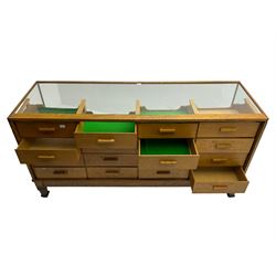 Early 20th century oak framed haberdashery shop counter, glass panel exterior enclosing sixteen graduating drawers with oak fronts and handles, raised on shaped tapering supports 