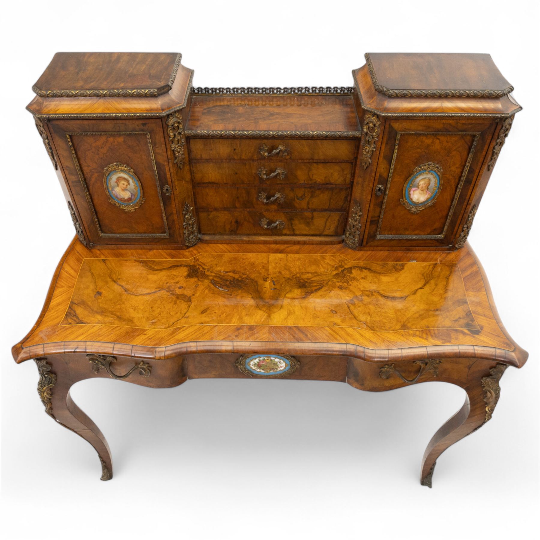 Late 19th-century Louis XV revival gilt bronze mounted and walnut Bonheur-de-Jour, rectangular-shaped serpentine form, with gilt bronze beading, frieze with two blind drawers and gilt bronze handles, centre drawer with large central serves style porcelain plaque, the elevated section with brass gallery above panelled cupboard doors also with serves style porcelain plaques and gilt bronze mounts, interior with single shelf, central section with four drawers each with gilt bronze handles, raised on slender cabriole legs fitted with gilt bronze rococo style masks and sabots