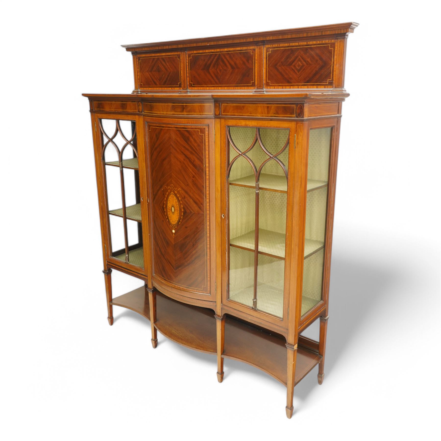 Maple & Co. Edwardian inlaid mahogany display cabinet, raised triple panelled back inlaid with quarter-matched veneers, break bow-fronted top over figured frieze with checkered stringing, enclosed by two astragal glazed doors and central panelled door, oval fan inlay with ribbon tie and trailing bell flowers, on square tapering supports with spade feet united by under tier, with 'Maple & Co.' circular makers plaque to the back