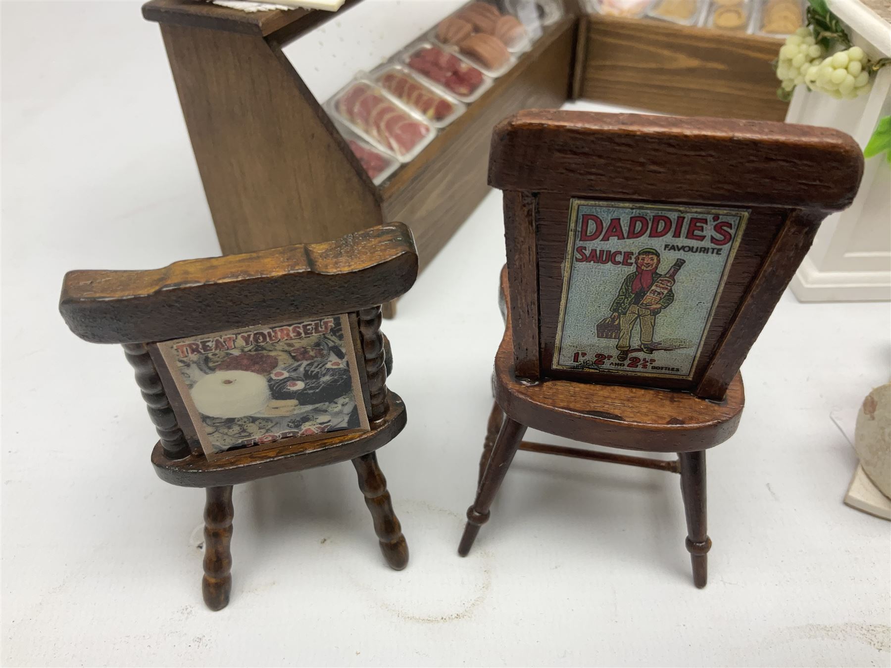 Collection of miniature dolls house grocery shop furniture and produce, comprising glazed corner counter with brass style cash register, shelved preserves unit, pies, cheese board and trays of meats, pair of white cheese counters, biscuit display with six tins, glazed preserves cabinet, pair of shop chairs and four further cheese boards complete with grapes and label flags