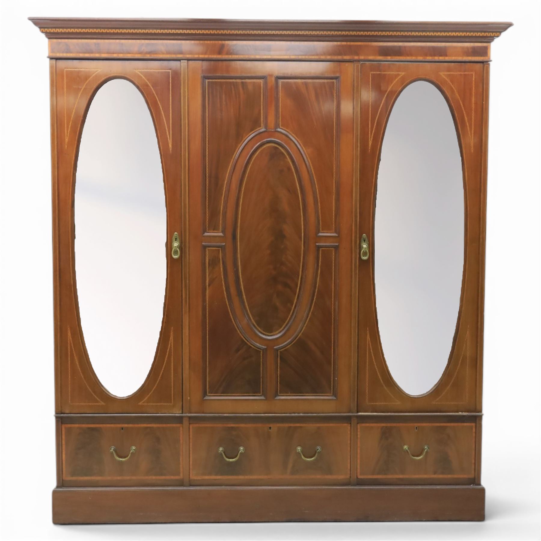 Edwardian inlaid mahogany triple wardrobe, featuring two oval mirror doors with bevelled plates, enclosing central figured veneered panel with mouled detail, above three base drawers