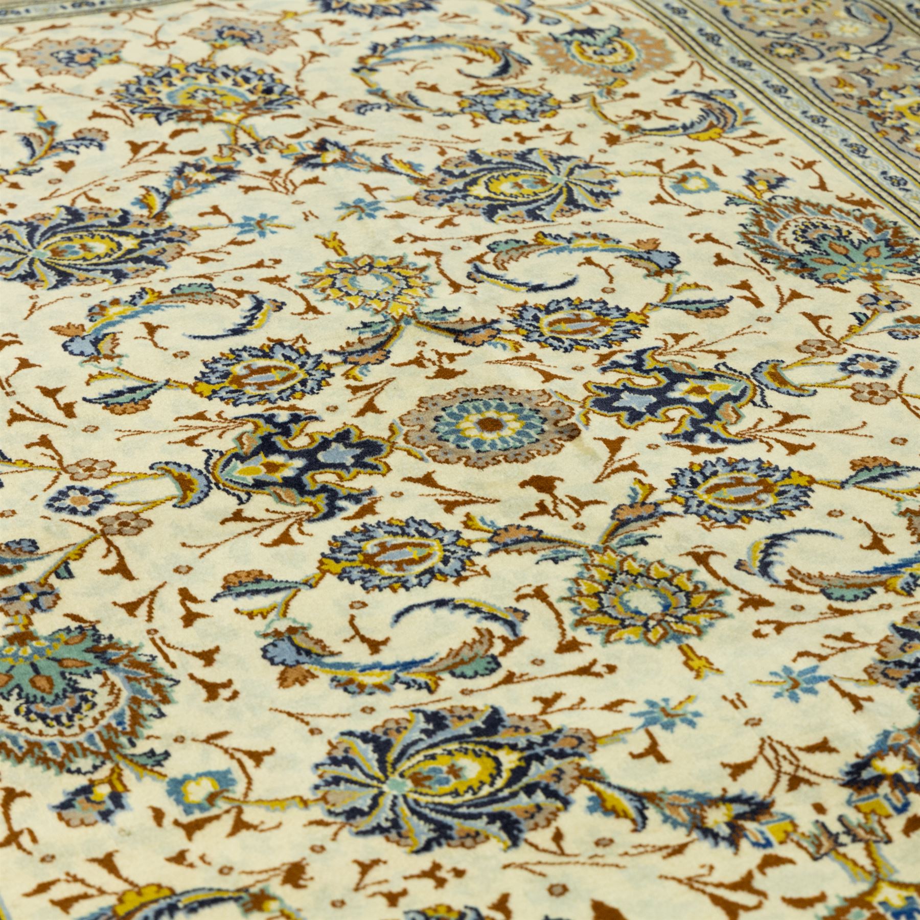 Central Persian Kashan hand-knotted carpet, ivory ground, with an all-over floral design of palmettes, scrolling vines and flowerheads in blue, ochre and beige tones, enclosed by a sage green border with repeating floral sprays and leafy scrolls, flanked by narrow guard stripes, with cotton fringes