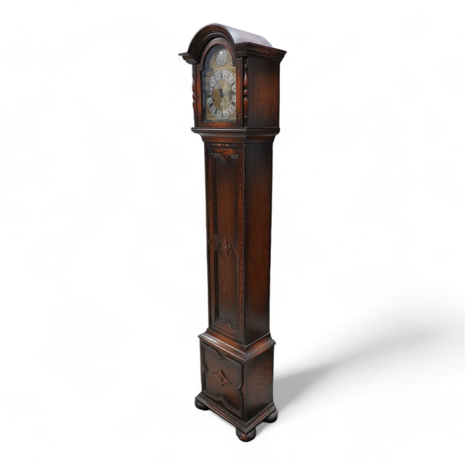 Oak cased 8-day grandmother clock c 1930 - with a broken arched pediment and break arch hood door flanked by turned pilasters, carved full length trunk door on a short plinth raised on bun feet, brass dial with cast spandrels, matted dial centre and silvered chapter with Roman numerals, three train spring driven movement chiming the quarters and hours on 8 gong rods.  With pendulum.