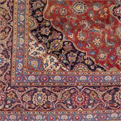 Central Persian Kashan hand-knotted carpet, madder red field with a central cusped medallion of ivory and deep indigo ground, enclosing a stylized floral rosette, framed by scrolling arabesques, field richly ornamented with an intricate network of flowering vinery, palmettes, and delicate floral sprays, spandrels in deep indigo, principal border of navy ground decorated with a continuous palmette and vine-scroll, flanked by multiple guard stripes, with cotton fringe ends 
